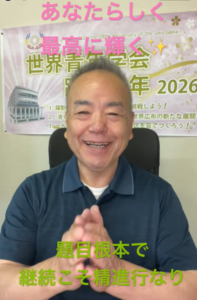 Smiling man in a blue polo sits in a chair with hands pressed together; banner with Japanese text and '2026' behind him.