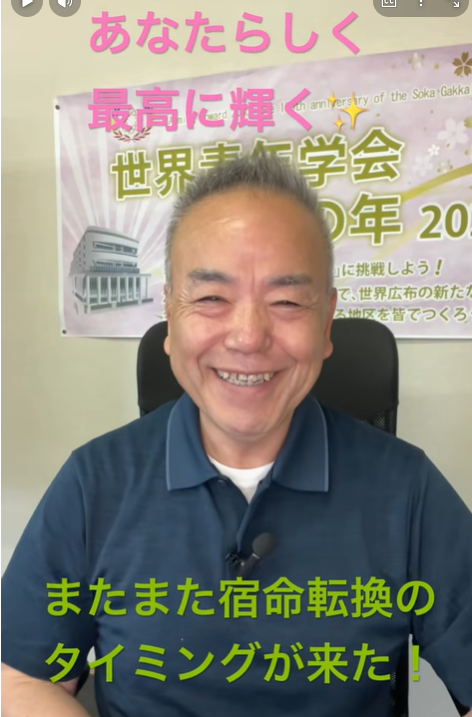 Smiling man in a blue polo shirt sits in front of a banner with pink and green Japanese text behind him, microphone clipped to his shirt