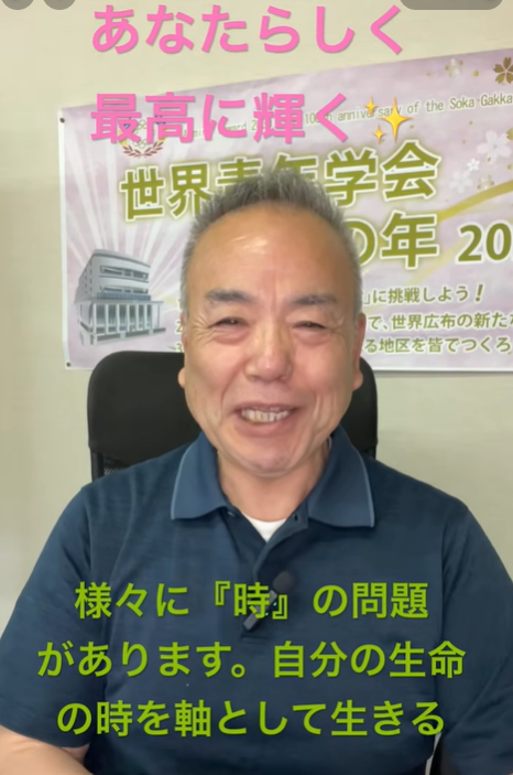 Smiling man in a blue polo shirt seated in front of a banner with Japanese text, the banner suggesting a conference or event backdrop.