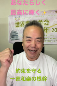 Smiling man in a white shirt raises a clenched fist, seated in front of a poster with Japanese text behind him; pink and green overlaid captions are visible.