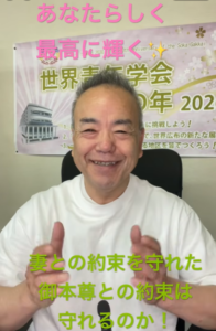 Smiling man in a white T-shirt giving thumbs up, seated in front of a wall banner with pink text above and green text across the bottom.