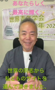 Smiling man in a blazer speaks to camera in a studio, with a poster behind him and large pink and green Japanese captions overlaid on the screen.