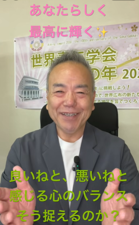 Smiling man in a suit speaks to the camera with a lapel microphone, standing in front of a banner with Japanese text.