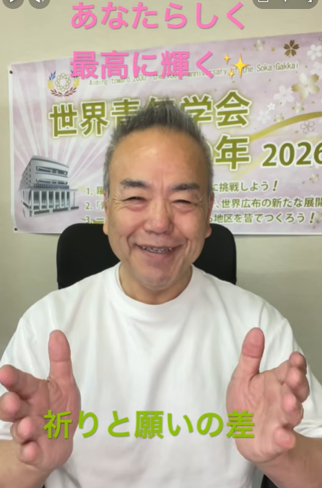 Smiling man in a white shirt sits in front of a pink-and-green banner with Japanese text; hands raised in a welcoming gesture.