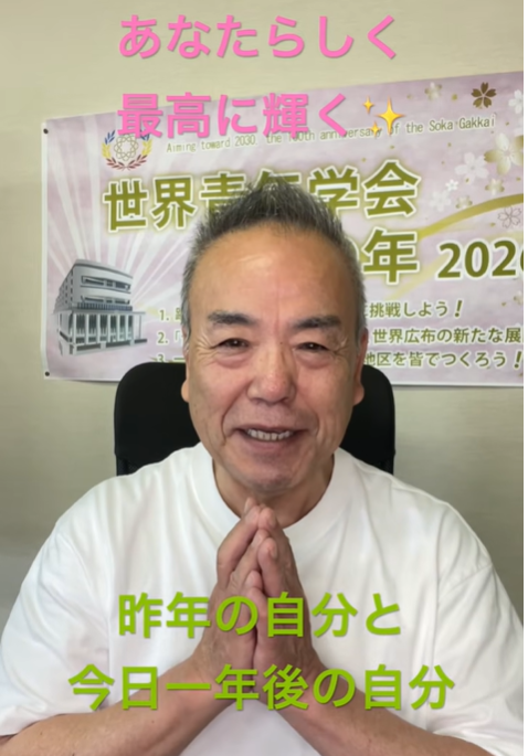 Smiling man in a white shirt with hands pressed together in a prayer-like gesture, seated before a banner with Japanese text behind him.