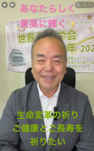Smiling man in a suit filming himself; background banner with Japanese text and pink/green captions across the screen.